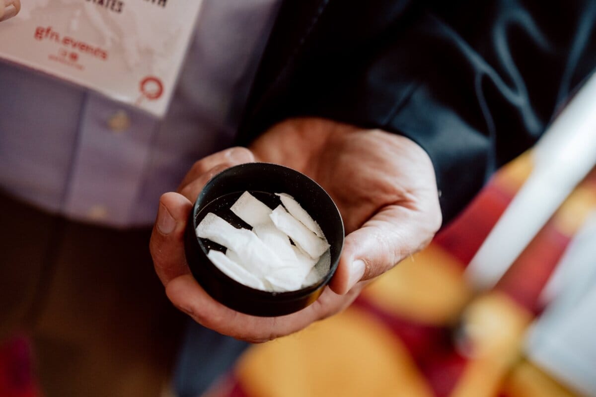 The person is holding a small black container filled with several white rectangular pads in his right hand. The person, presumably an event photographer, is wearing a dark jacket and light blue shirt, and part of a lanyard with a badge can be seen around his neck. 