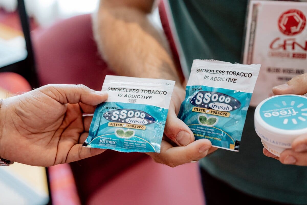   Close-up of two hands holding bags of smokeless tobacco with the label "S0000 Fresh Filter Tobacco" and the text "Smokeless tobacco is addictive." The other hand holds a container with the label partially visible in the background, capturing the moment like an event photographer documenting an event. 