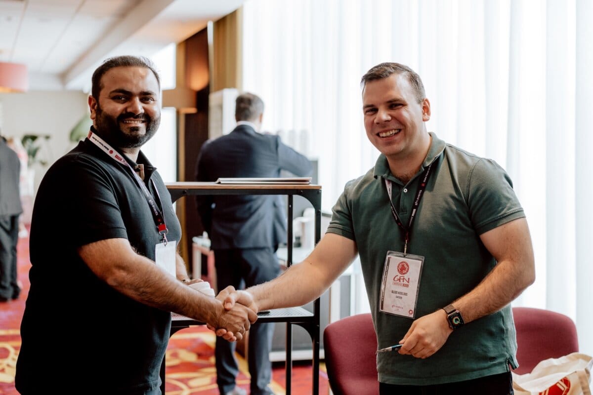 Two men smile and shake hands at an event taking place in a room. Both are wearing conference badges. The man on the left is wearing a black polo shirt, and the man on the right is wearing a green polo shirt. They are standing in front of a white curtain, with other attendees in the background, which was perfectly captured by our event photographer.   