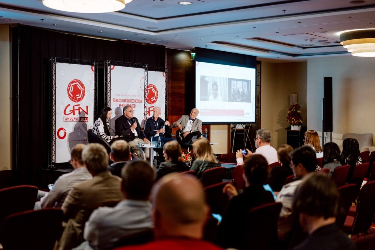 A group of people sit on a panel in front of the audience during the conference. Behind them, banners with logos and a screen displaying a video call set the stage for a photo essay of the conference. Participants sit facing the stage, listening and taking notes in what resembles a conference room.  