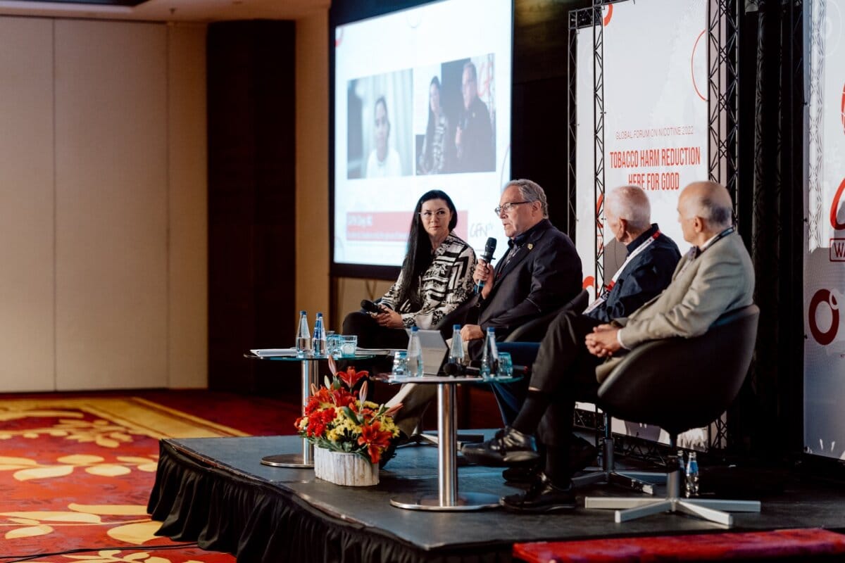 Four people sit on a stage panel and discuss. One person speaks into a microphone while the others listen attentively. A screen in the background shows additional participants joining in virtually. The stage shown in the photo essay of the conference is decorated with flowers and event branding.   