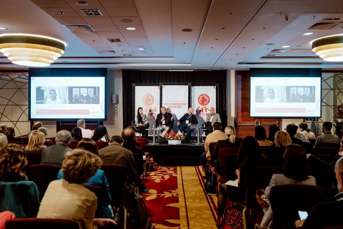 A conference room filled with attendees observing a panel discussion. Four panelists sit on stage with microphones and lead the conversation. Two large screens display the event's branding and visuals, with a prominent banner with the organization's logo behind them, perfect for photo coverage of the conference.  