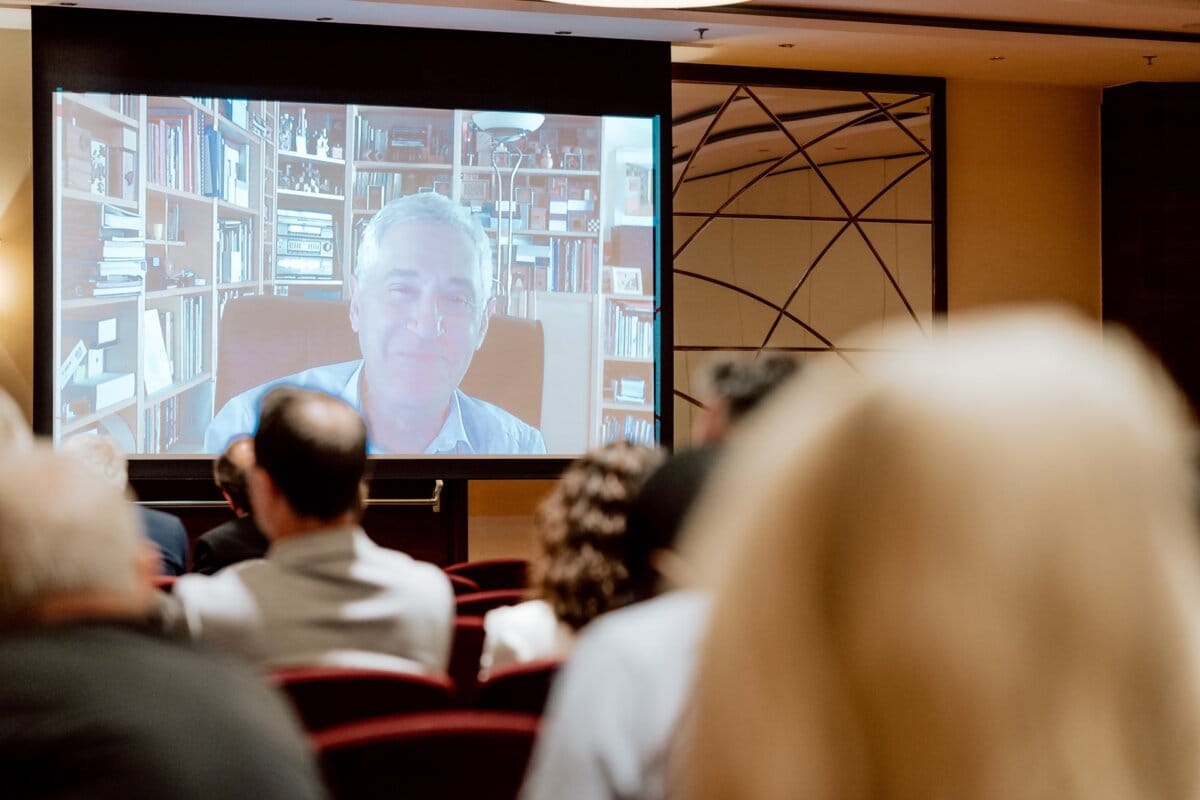 A group of people attending an event, sitting opposite a large screen displaying live video of a man speaking. The man is in a room with shelves filled with books in the background. This photo essay of the conference shows the attentive audience and the speaker's insightful presentation.  