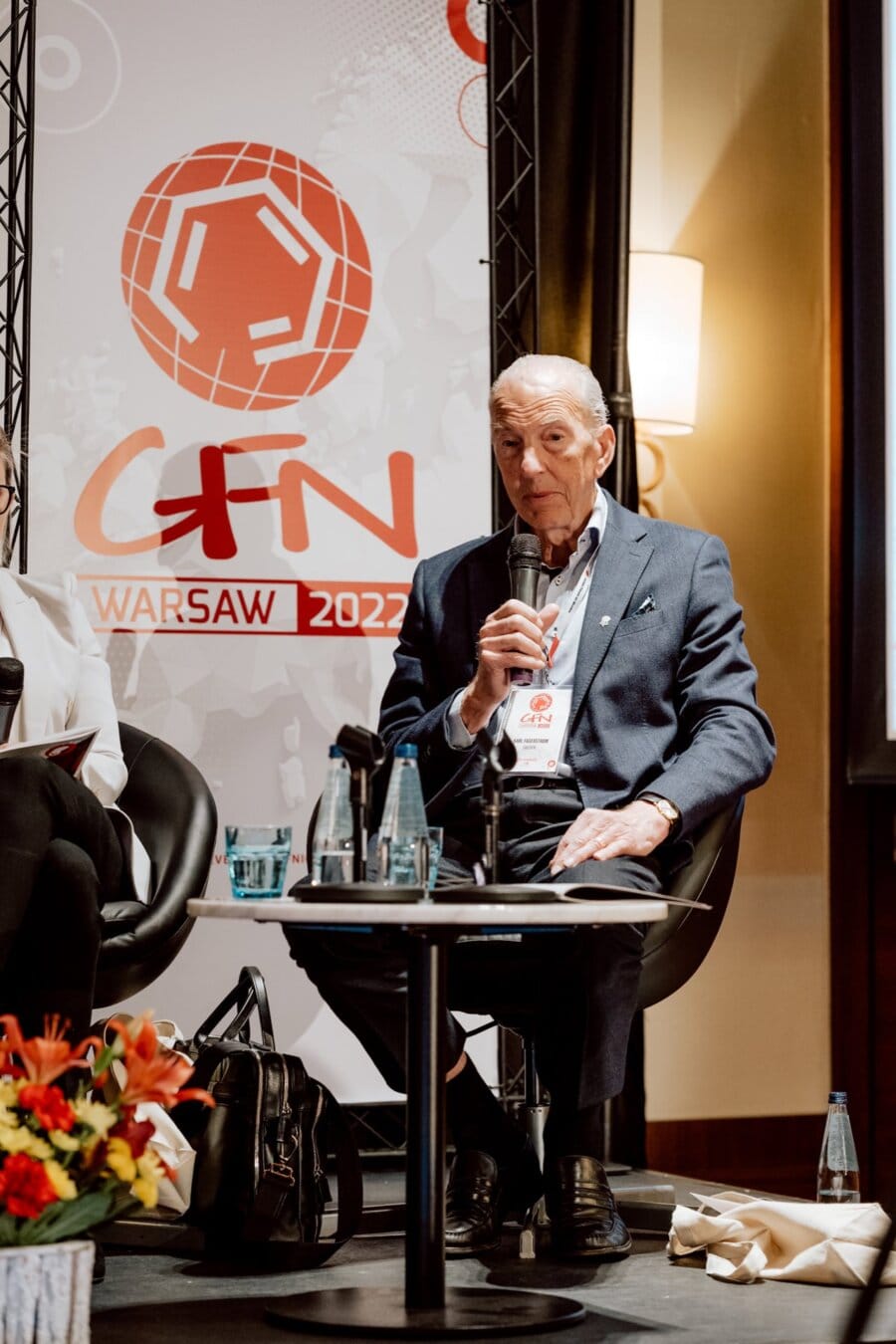 An older man in a blue jacket sits at the conference and speaks into a microphone. Behind him is a banner with the words "GFN WARSAW 2022" and various designs. On the table in front of him is a bouquet of flowers and bottles of water, highlighting the details of the conference photo-op.  