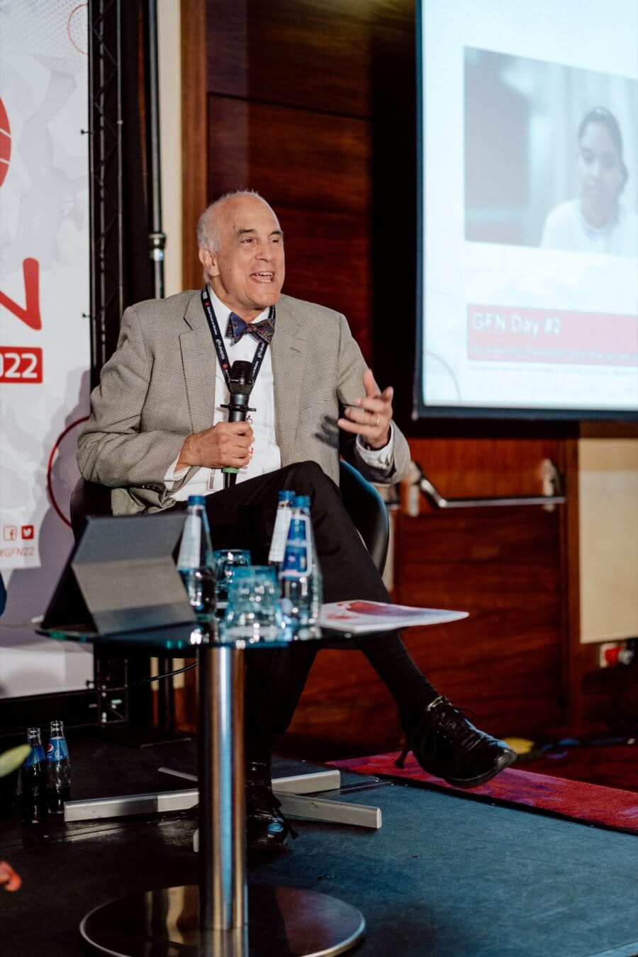 A man in a gray jacket and bow tie speaks into a microphone, sitting in a black chair on the stage. Behind him, a projector screen displays a virtual meeting. On a small table in front are two bottles of water and a glass of water, giving the scene the feel of an engaging photo essay of a conference.  