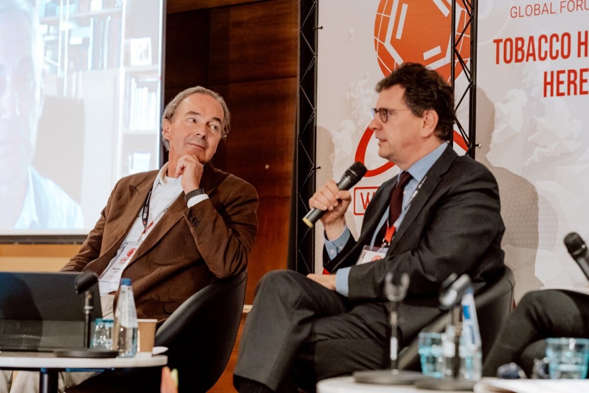 Two men sit on stage during a panel discussion. The man on the right is speaking into a microphone, wearing glasses and a suit. The man on the left, wearing a brown jacket, looks at him intently. A photo of the conference reveals event signage in the background referring to the World Forum on Tobacco Harm Reduction.   