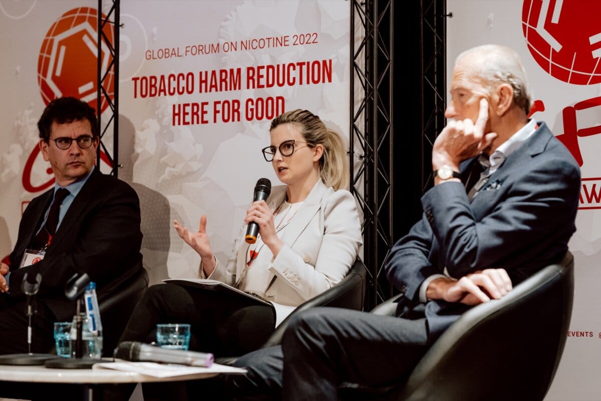 Three people sit on the stage of the World Forum on Nicotine 2022 in front of a banner that reads "We will reduce the harms of smoking for good." The woman in the middle speaks into a microphone, while two men on either side listen intently, creating an engaging report on the event. 