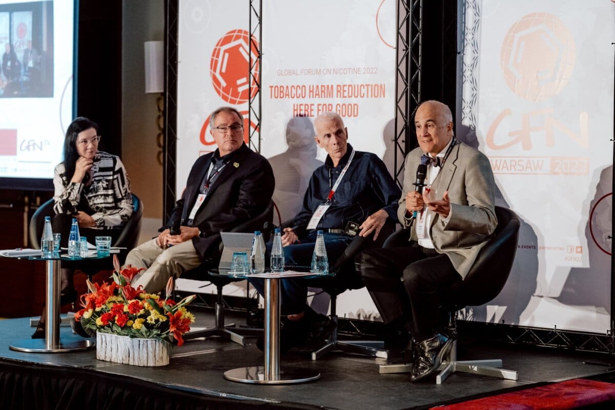 A panel of four people, three men and one woman, sitting on stage at the "World Nicotine Forum 2022" in Warsaw. Behind them is a banner that reads "Reducing tobacco-related harm: here for good." One man speaks into a microphone. The front is decorated with a flower arrangement. This photo report from the event poignantly shows their discussion.    