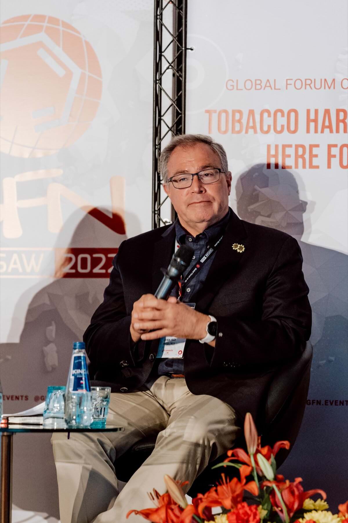 Mężczyzna siedzi na scenie podczas Światowego Forum omawiającego redukcję szkód spowodowanych paleniem tytoniu. Trzyma mikrofon, ma na sobie okulary, ciemną kurtkę i beżowe spodnie. Na stole obok niego stoją dwie butelki z wodą. Tło zawiera loga forum i tekst, tworząc idealną fotorelację z wydarzenia.