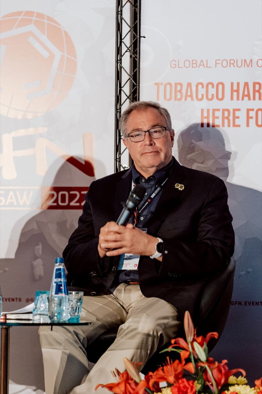 A man sits on stage at a World Forum discussing tobacco harm reduction. He is holding a microphone, wearing glasses, a dark jacket and beige pants. On a table next to him are two bottles of water. The background includes the forum's logos and text, creating a perfect photo-essay of the event.   