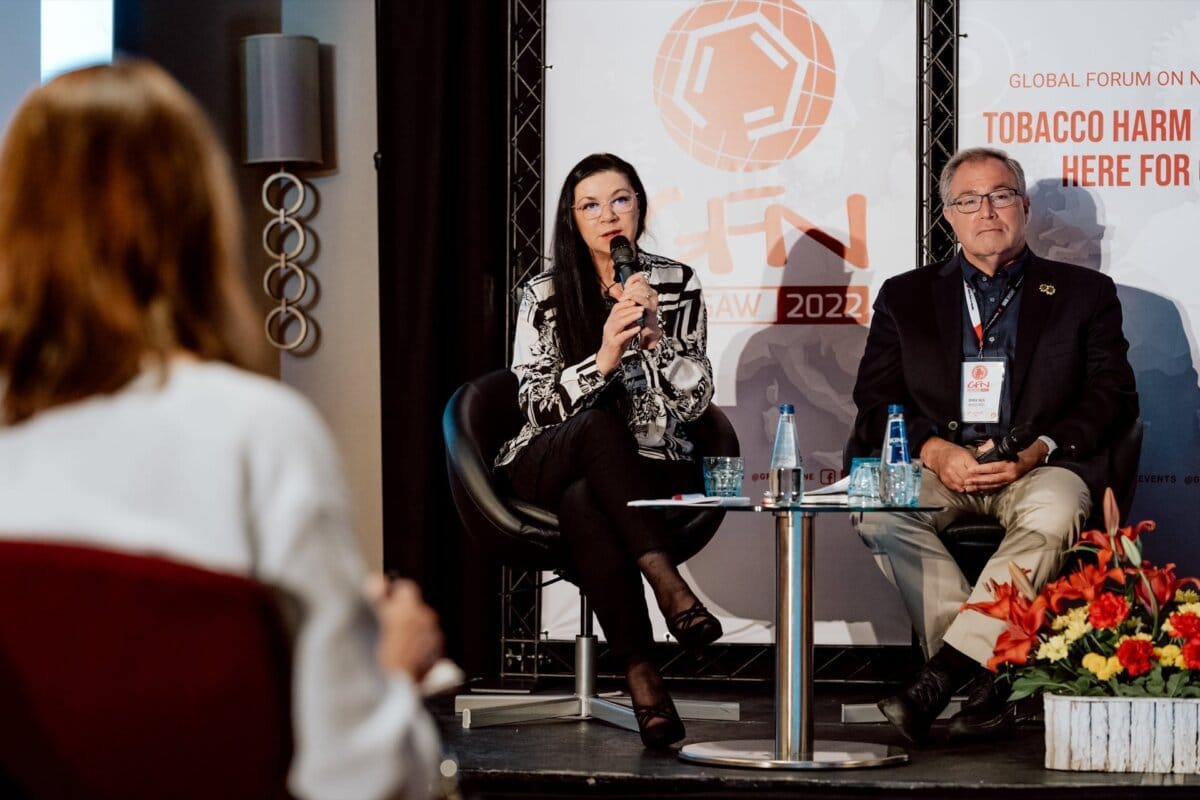 Three people take part in a panel discussion at the World Forum on Nicotine 2022. The photographer captured the woman on the left speaking into the microphone, while the man on her right sits attentively. The two are standing on stage with a banner behind them. Another person's back is visible in the foreground, adding depth to the conference photo.   