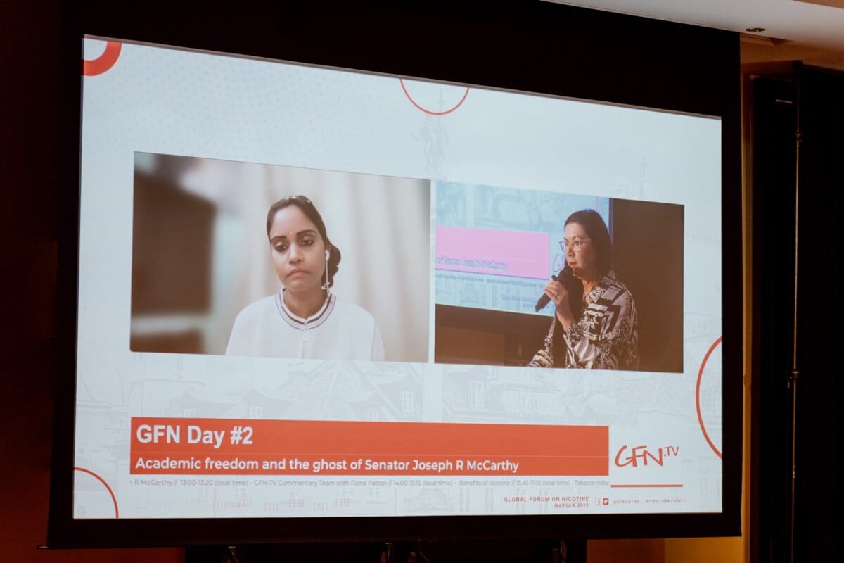 The large screen shows two speakers participating in a discussion. The left speaker, a woman, appears thoughtful while the right speaker speaks into the microphone. Below is a banner with the caption "GFN Day #2, Academic Freedom and the Spirit of Senator Joseph R. McCarthy," which captures the essence of this photo essay from the conference.  