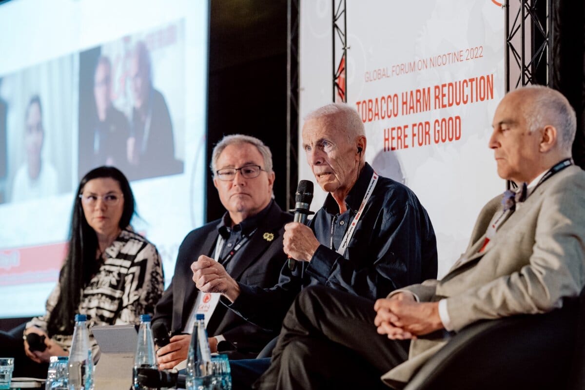 Four people sit and speak on a panel discussion at the "Global Forum on Nicotine 2022." The background emphasizes the theme "Here for good tobacco harm reduction." This photo report from the conference shows three men and one woman on stage, with microphones and water bottles on a table in front of them.  