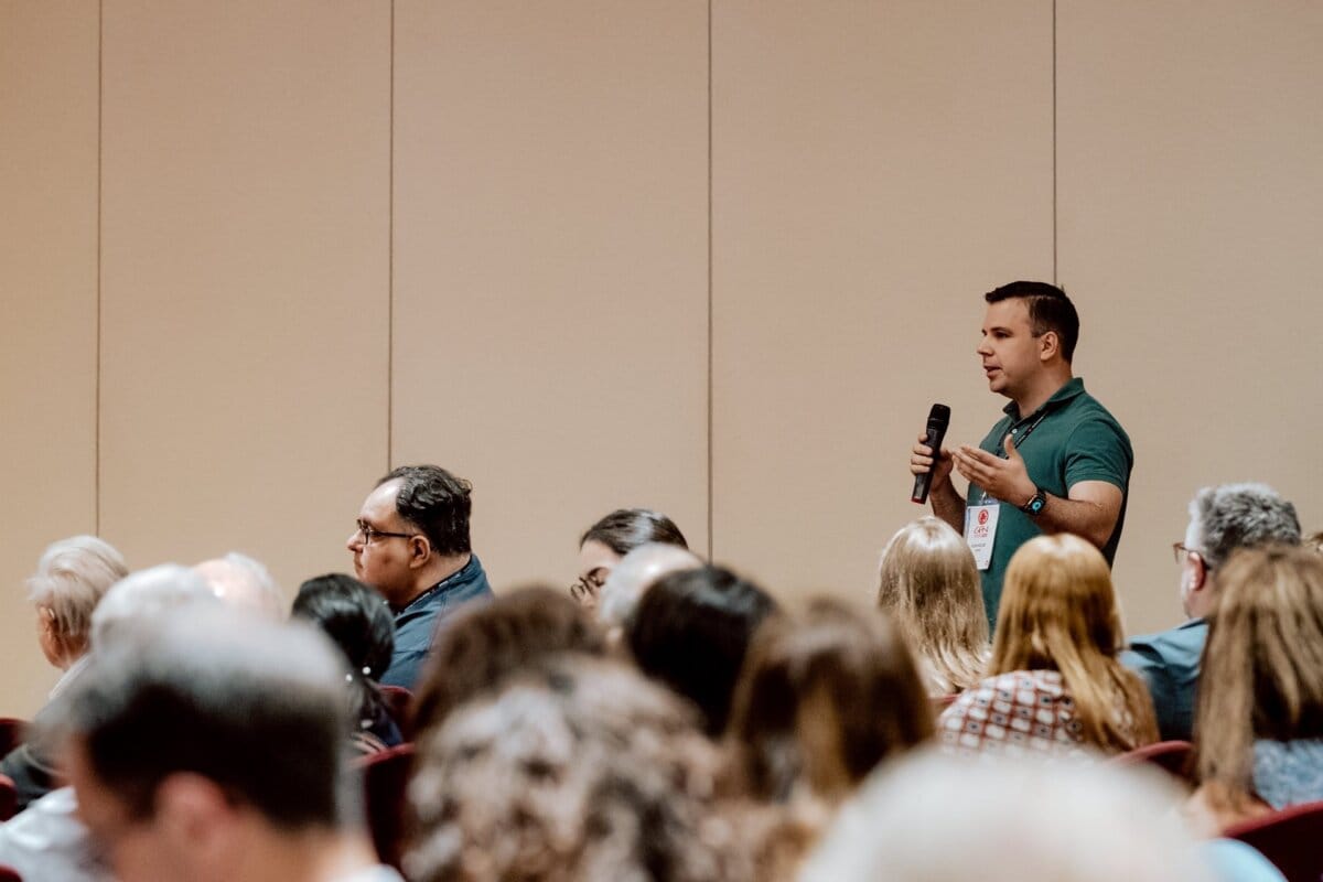 A man stands with a microphone and speaks to an audience in a conference room, his green shirt glowing against the beige walls. Engaged in discussion, he captivates the attentive listening audience. This photo report from the event captures the essence of their engagement and interaction.  