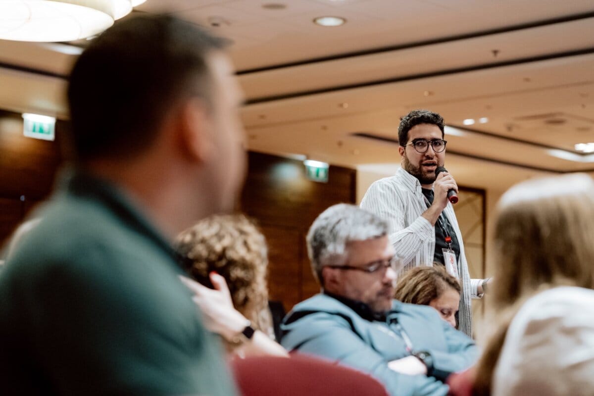 A man standing and holding a microphone is speaking to an audience in a conference room. People sit and listen attentively, while various people can be seen in the foreground. The room has warm lighting and a professional atmosphere, perfectly captured in this photo report of the event.  