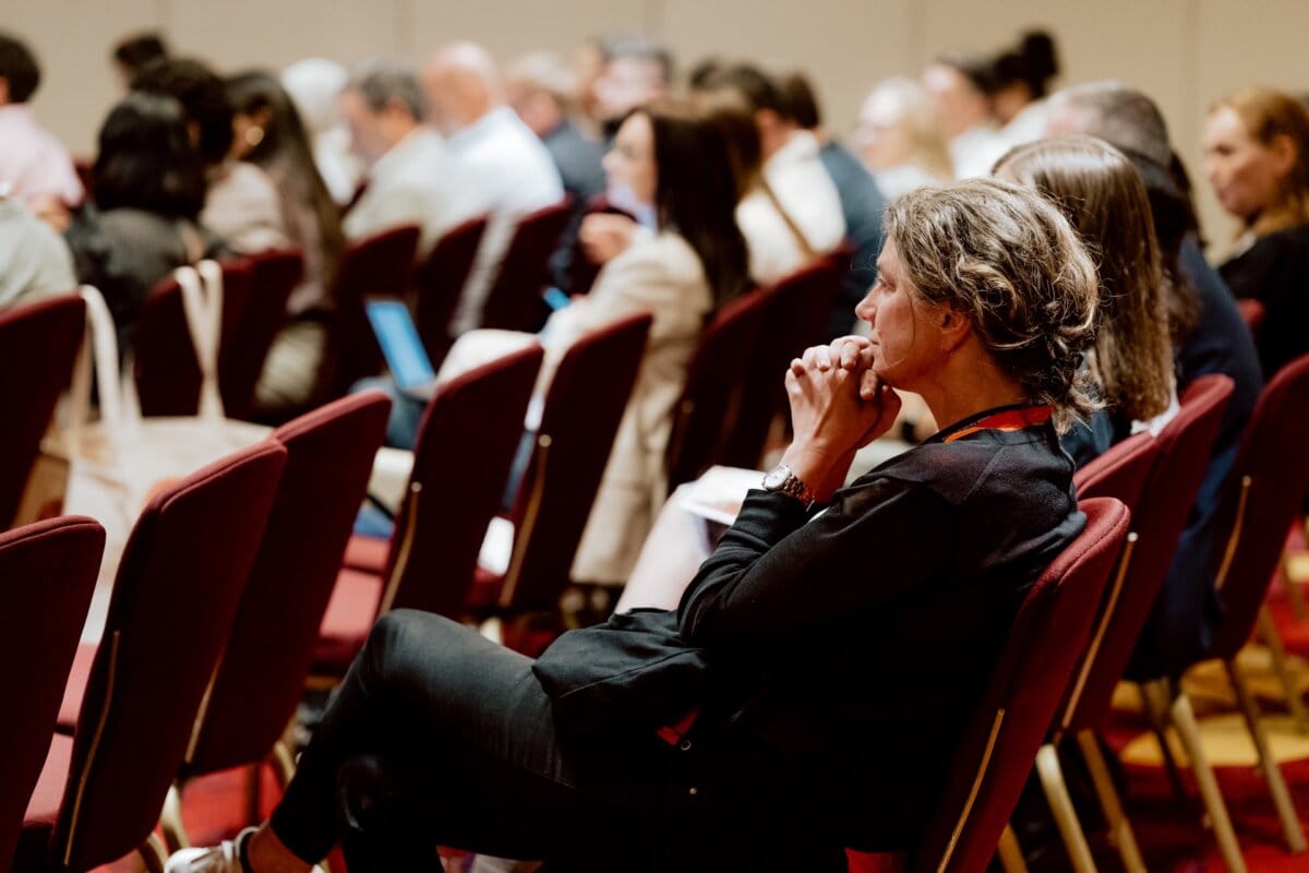 A woman with gray hair, sitting in a row of chairs with red cushions, appears to be immersed in her thoughts, with her hands clasped in front of her face. She is part of an audience attending a closed-door conference, captured as part of a photo essay of the conference, which shows many people lined up in rows of chairs. 