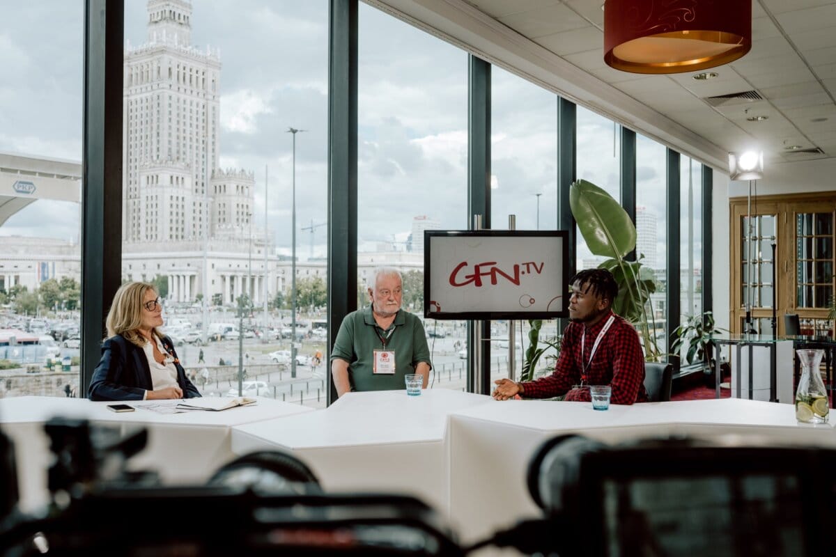 Three people are seated around a table in a well-lit studio with large windows. A TV screen with the words "CFN TV" is visible in the background. Outside the windows one can see the city skyline with a distinctive tall building. Photographic equipment in the foreground captures this dynamic snapshot of the event.   