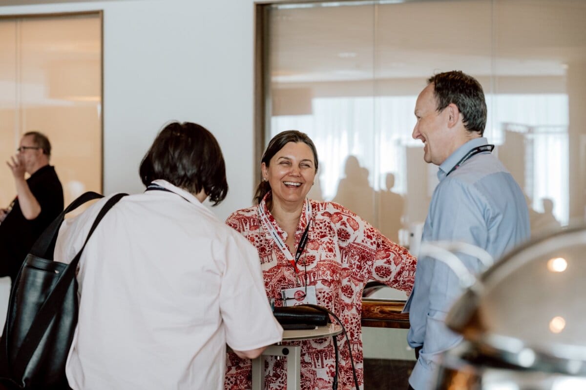 Three adults, two women and one man, are standing and having a cheerful conversation in the room. The woman in the middle smiles warmly. They are wearing conference badges, suggesting they may be at a professional or networking event - perhaps captured for a photo essay of the event.  