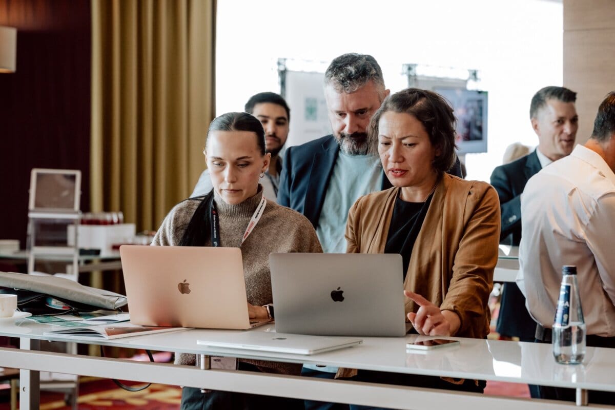 A group of people stand around a tall table with two MacBook laptops. Two women at the front are focused on the screens, while a man stands behind them and watches. Other people in business attire can be seen in the background, engrossed in conversation as part of a photo essay on the conference.  