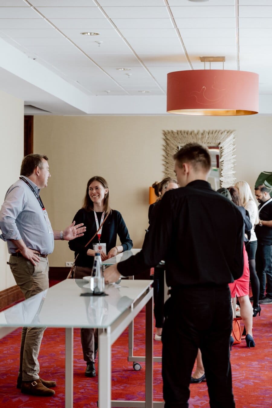 A group of people are talking in a conference room with white walls and a red carpet. Some are standing at a long white table with a bottle of water, while others are talking at a wall mirror. The atmosphere seems casual and lively, perfect for a photo essay on the conference.  