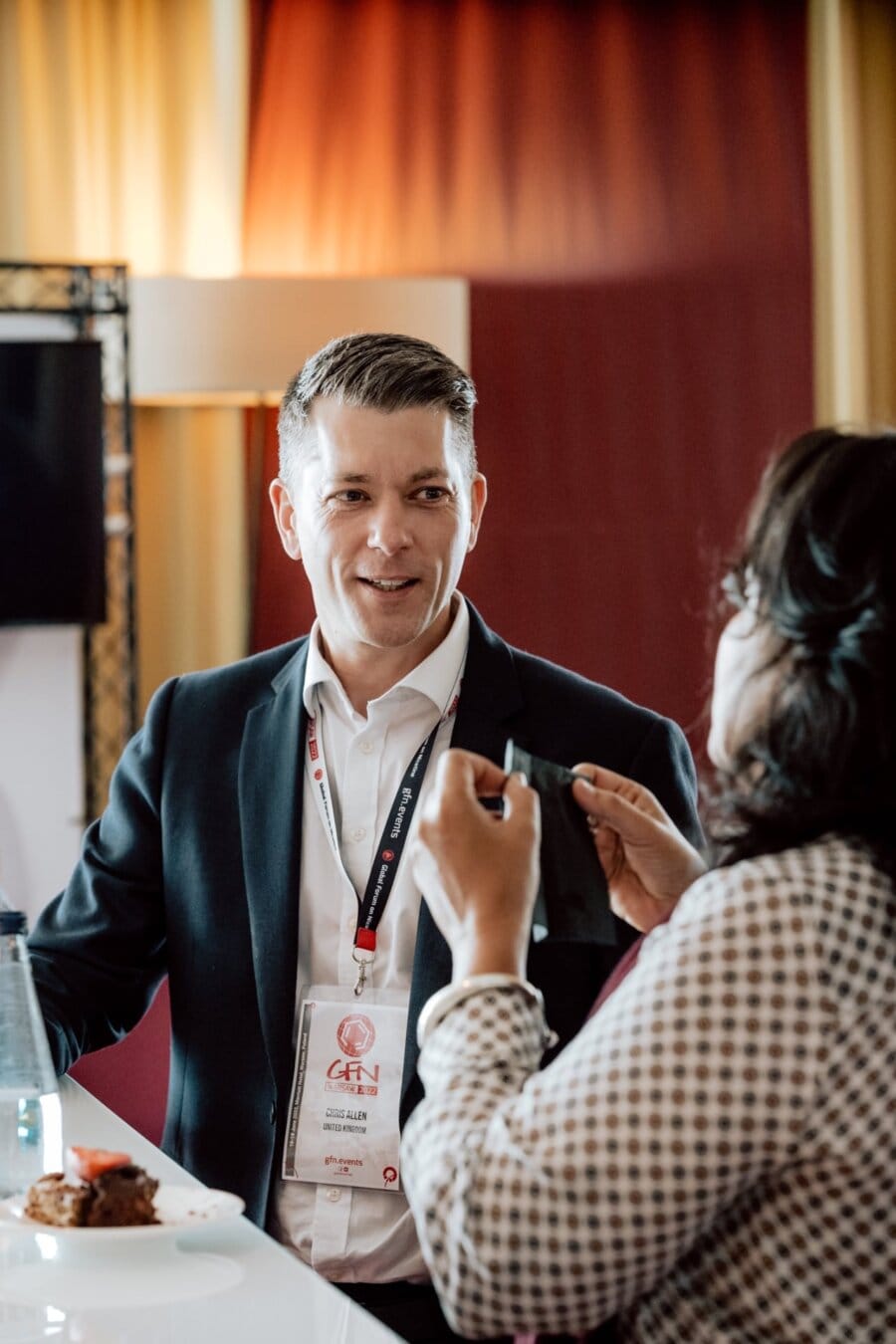 A man in a suit and conference badge is standing at a white counter and talking to a woman with her back turned to the camera. Captured by an event photographer, they are in a well-lit room, perhaps at a conference or professional event. 