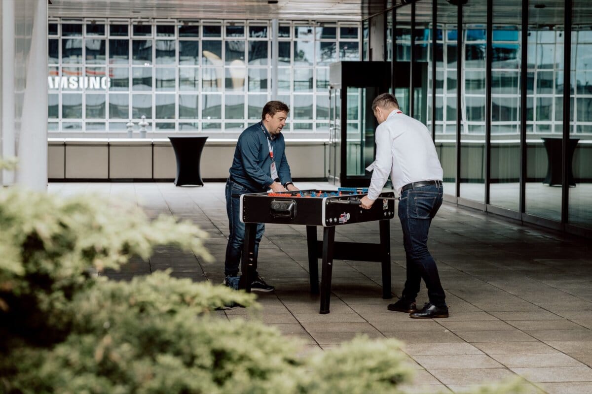 Two people play foosball on an outdoor patio with glass walls and city buildings in the background. The patio floor is lined with large stone slabs, and a small shrub is visible in the foreground, adding to the charm of this casual moment worthy of an event photo shoot. 