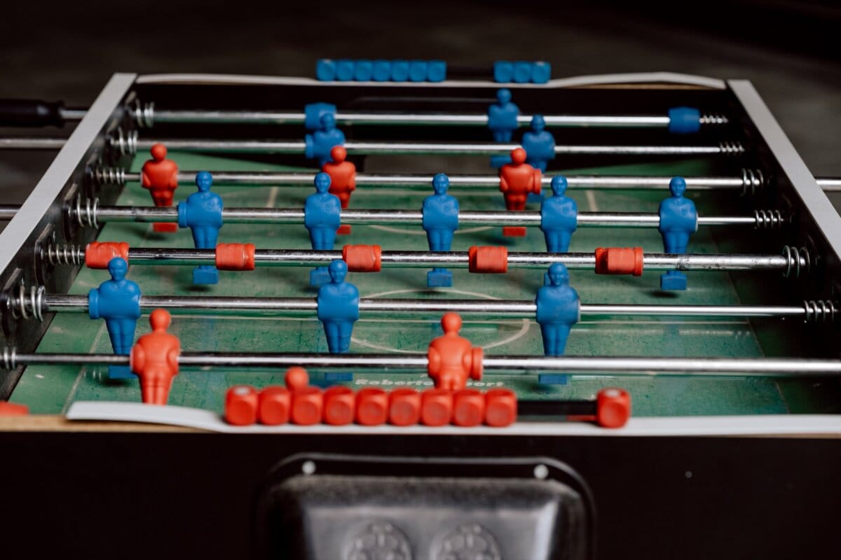 A photo taken by the event's photographer shows a foosball table with blue and red player figurines. The table consists of rows of figurines connected by metal rods, ready to play. The blue figures are positioned for defense and offense, while the red figures are arranged in a similar fashion, all on a green playing surface.  