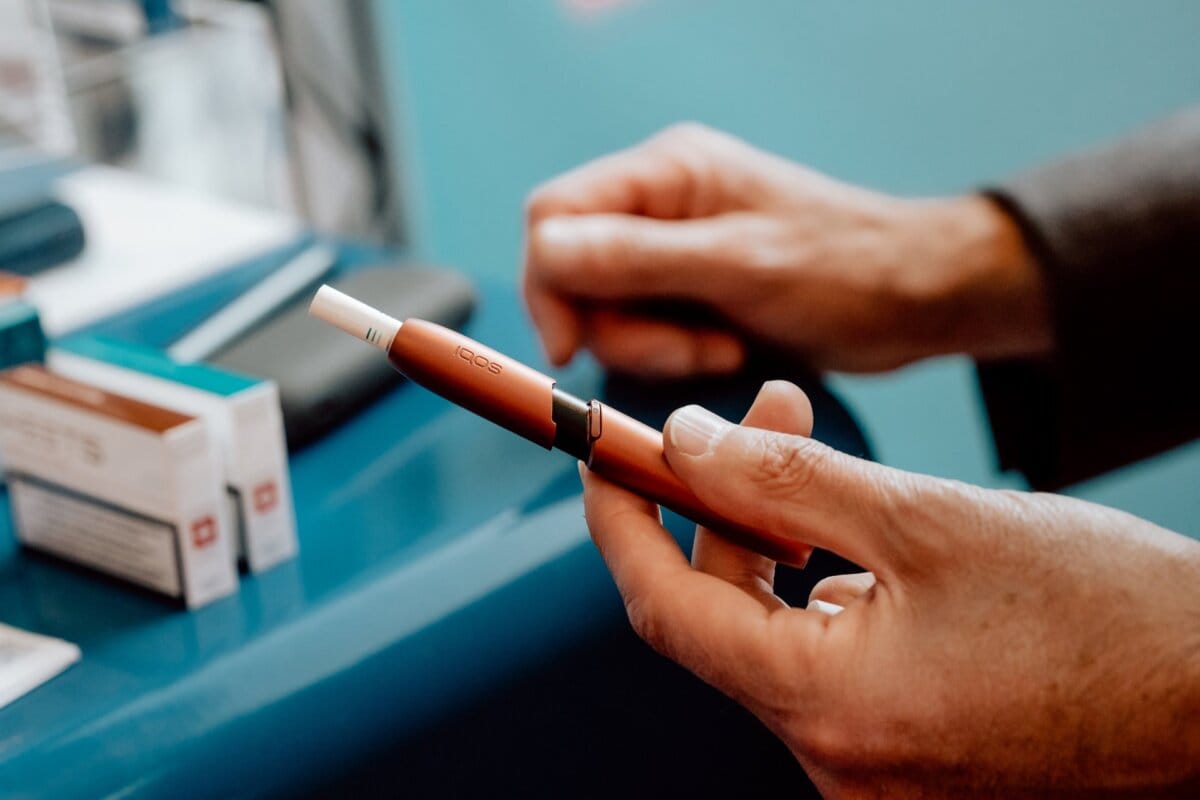 A close-up of the hand of a person holding an electronic cigarette. The e-cigarette is sleek, with a brown finish and a white tip. There are boxes and a blue surface in the background, suggesting a retail or display setting, perhaps part of an offotore from an event. The focus is on the device.   