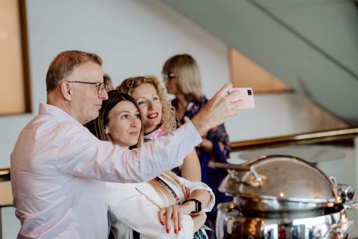 Three people, two women and one man, stand close together and pose for a selfie. The man is holding a pink smartphone to capture the moment. Dressed in semi-formal attire, they give the impression of attending a modern event held indoors, with a metal serving platter nearby, perhaps to photo-shop the event.  