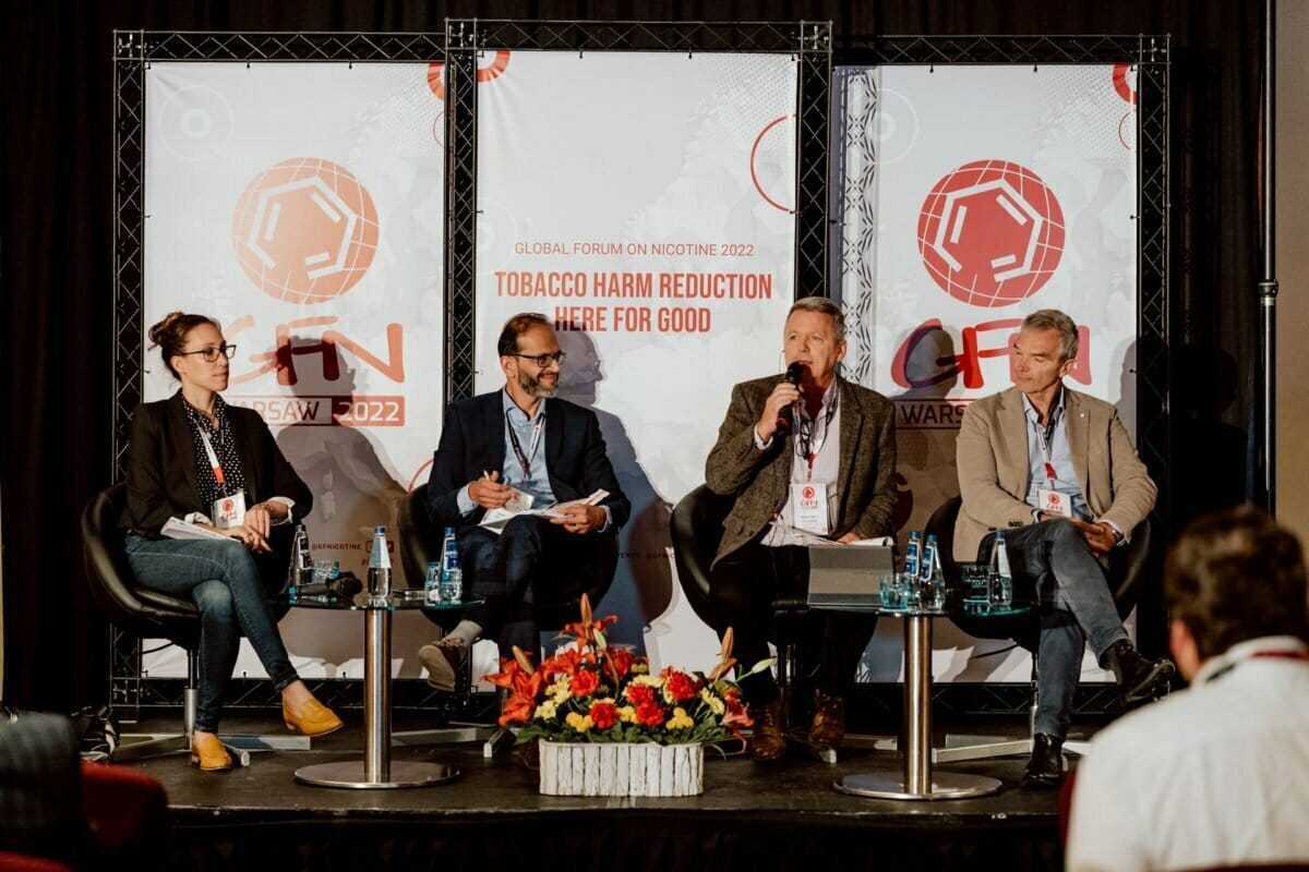 A panel of four sitting on chairs on a stage take part in a discussion at the World Nicotine Forum 2022 in Warsaw, Poland. In the background is the logo of the event and the slogan "Here for good tobacco harm reduction". This striking photo report from the event shows part of the audience partially visible.  