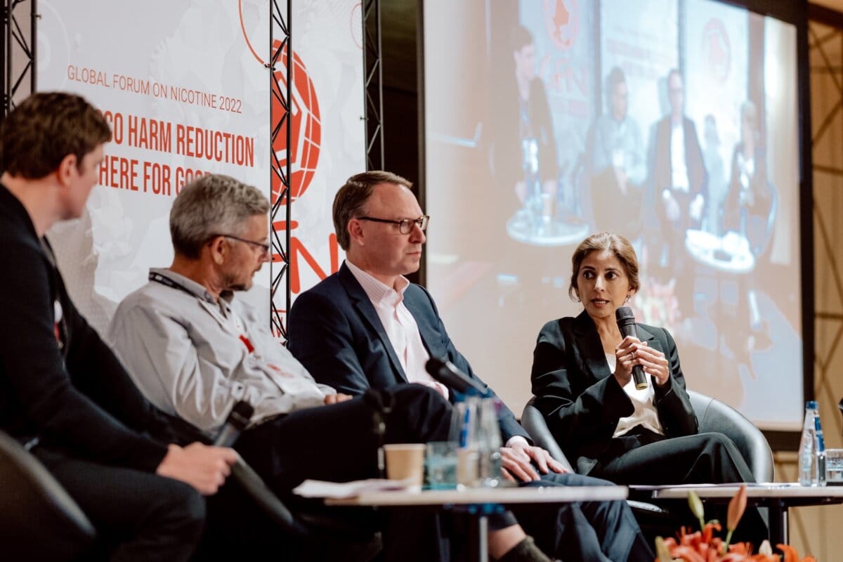 Four people are sitting on the stage and taking part in a panel discussion as part of the "Global Forum on Nicotine 2022." One woman is speaking, holding a microphone. Behind them, a large screen displays an enlarged image of the discussion, captured as part of a detailed reportage of the event.  