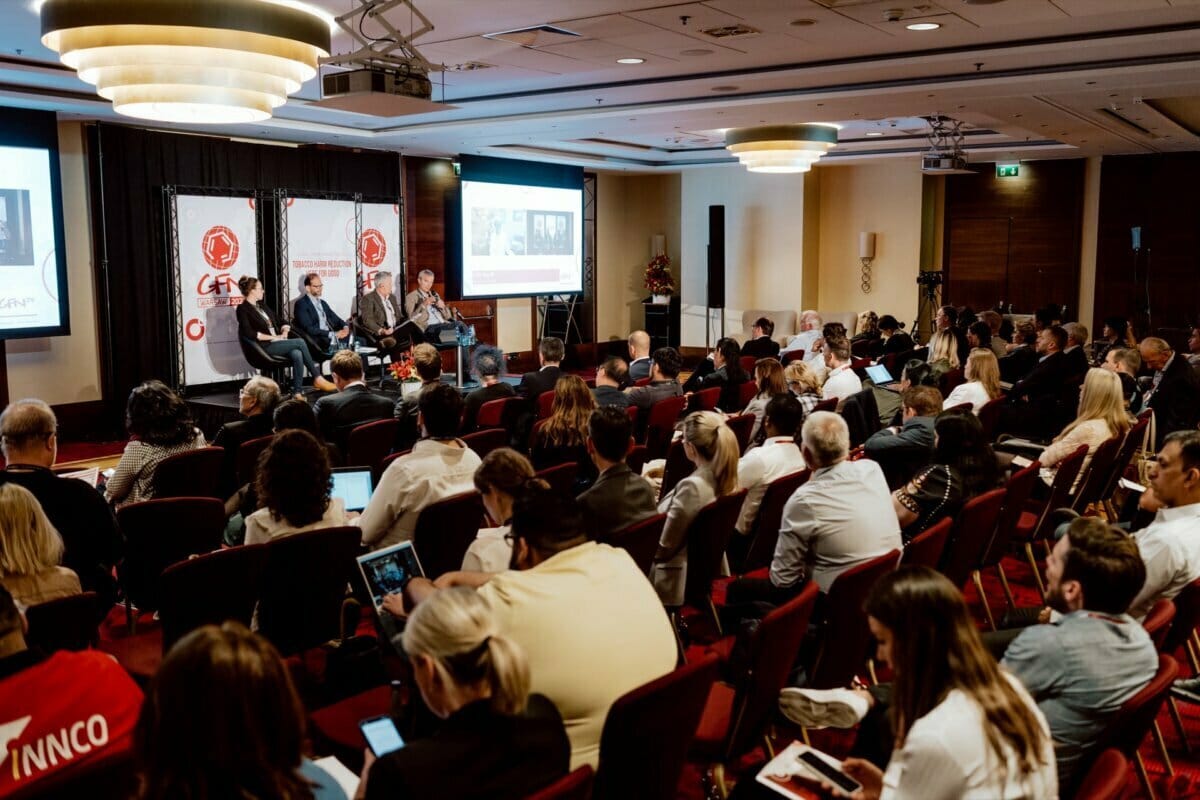 The conference room filled with seated attendees is opposite the stage. Four speakers sit on a panel, immersed in discussion, while a presentation is projected behind them. People are taking notes on laptops and listening intently. Red badges and conference banners are visible, capturing the essence of the event's photo coverage.   
