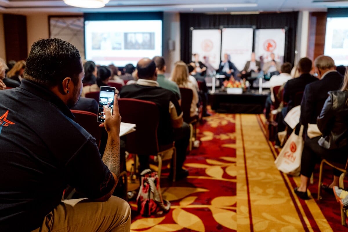 A man in the foreground picks up his phone to take a photo or video during a conference. Participants are seated facing the stage, where a panel of speakers is engaged in discussion. The room is well-lit and the presentation slides are displayed on screens behind the speakers, which is perfect for photo coverage of the event.  