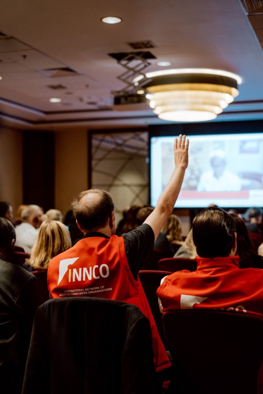 A man in a red vest sits with his back to the camera and raises his hand in a dimly lit conference room. He faces a large screen displaying the presentation while other participants, some also wearing red vests, sit around him. A modern ceiling fixture adds to the atmosphere of this photo essay of the event.  