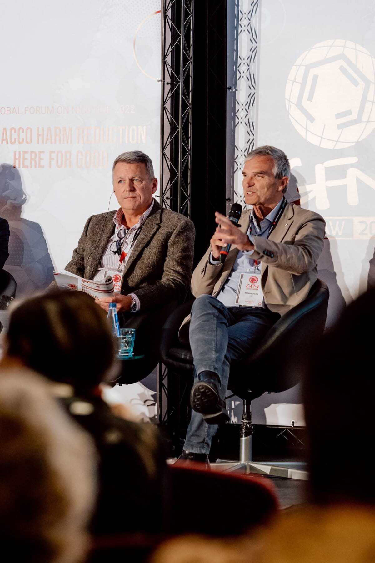 Podczas konferencji na panelu scenicznym siedzi dwóch mężczyzn. Mężczyzna po lewej stronie trzyma książeczkę, drugi przemawia za pomocą mikrofonu. Tło za nimi wyświetla logo i tekst. Oboje wydają się zaangażowani w tę fotorelację z konferencji.