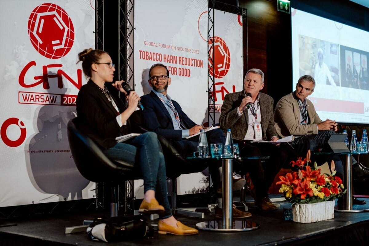 A panel discussion at the World Nicotine Forum 2022 in Warsaw, Poland, captured in a photo report from the event. Four speakers, three men in suits and a woman, sit on stage with microphones. Banners behind them proclaim: "Good tobacco harm reduction." The table is decorated with flower arrangements and water bottles.   