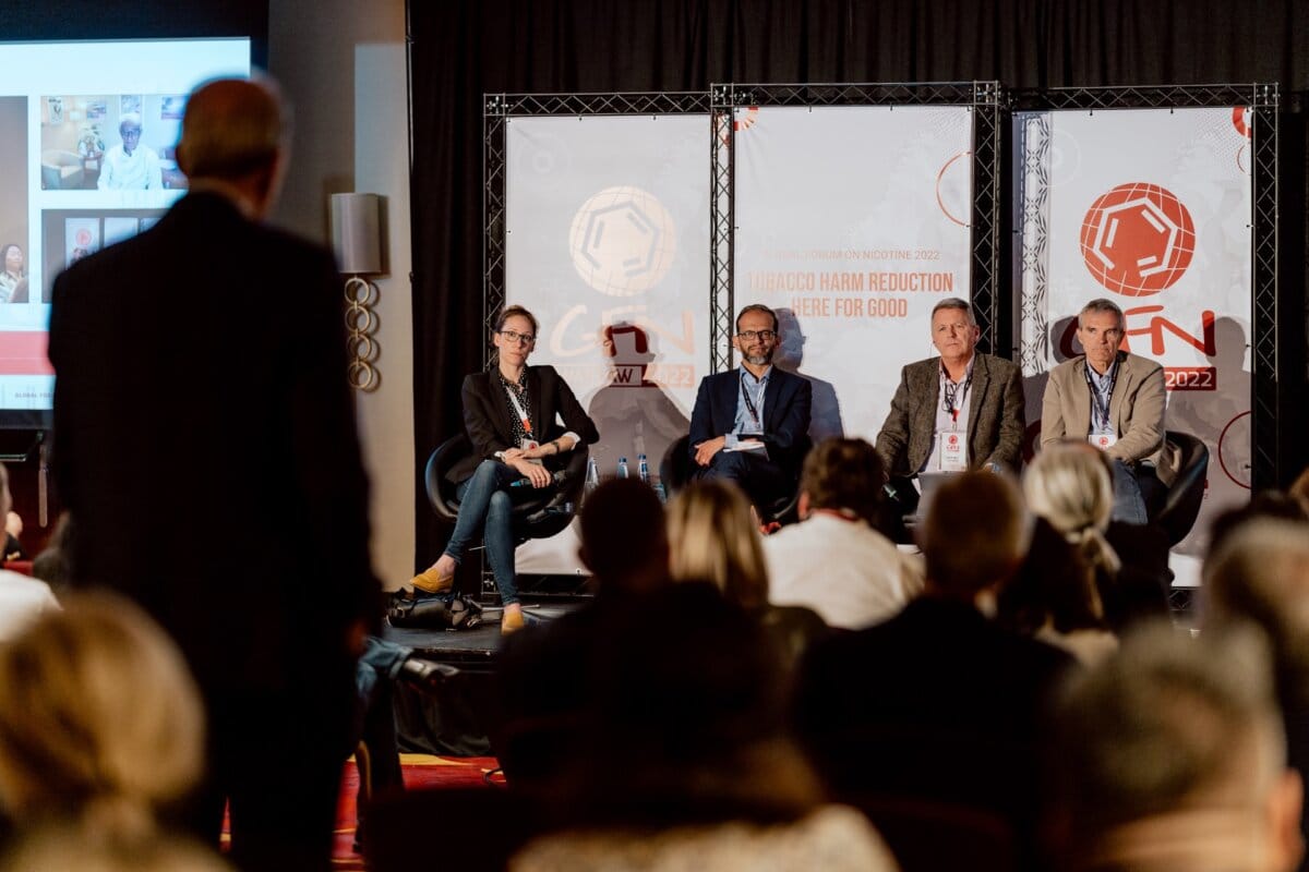 A group of five people taking part in a panel discussion are sitting on the stage. The audience can be seen in the foreground. Behind the panelists hang banners with the words "REDUCING THE DESTRUCTIVE DAMAGE of Tobacco 2022" and "TOGETHER FOR GOOD." The room is dimly lit, focused on the stage, perfectly captured by our event photographer.   