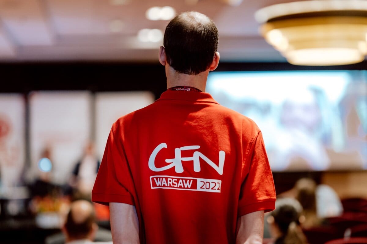 A person wearing a red shirt with "GFN WARSAW 2022" printed on the back faces a blurry conference room filled with attendees and a large screen displaying a presentation. The room was lit with natural and artificial light, capturing the essence of the conference photo essay. 