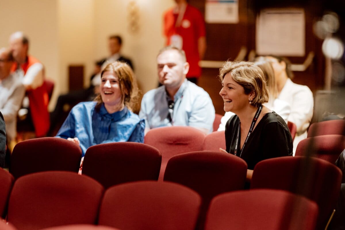 A group of people sitting on red auditorium chairs, smiling and engaged. A woman in a blue shirt laughs with a man in a gray shirt and a woman in a black top. Other people sit in the background, highlighting the lively atmosphere captured by the event's photographer.  