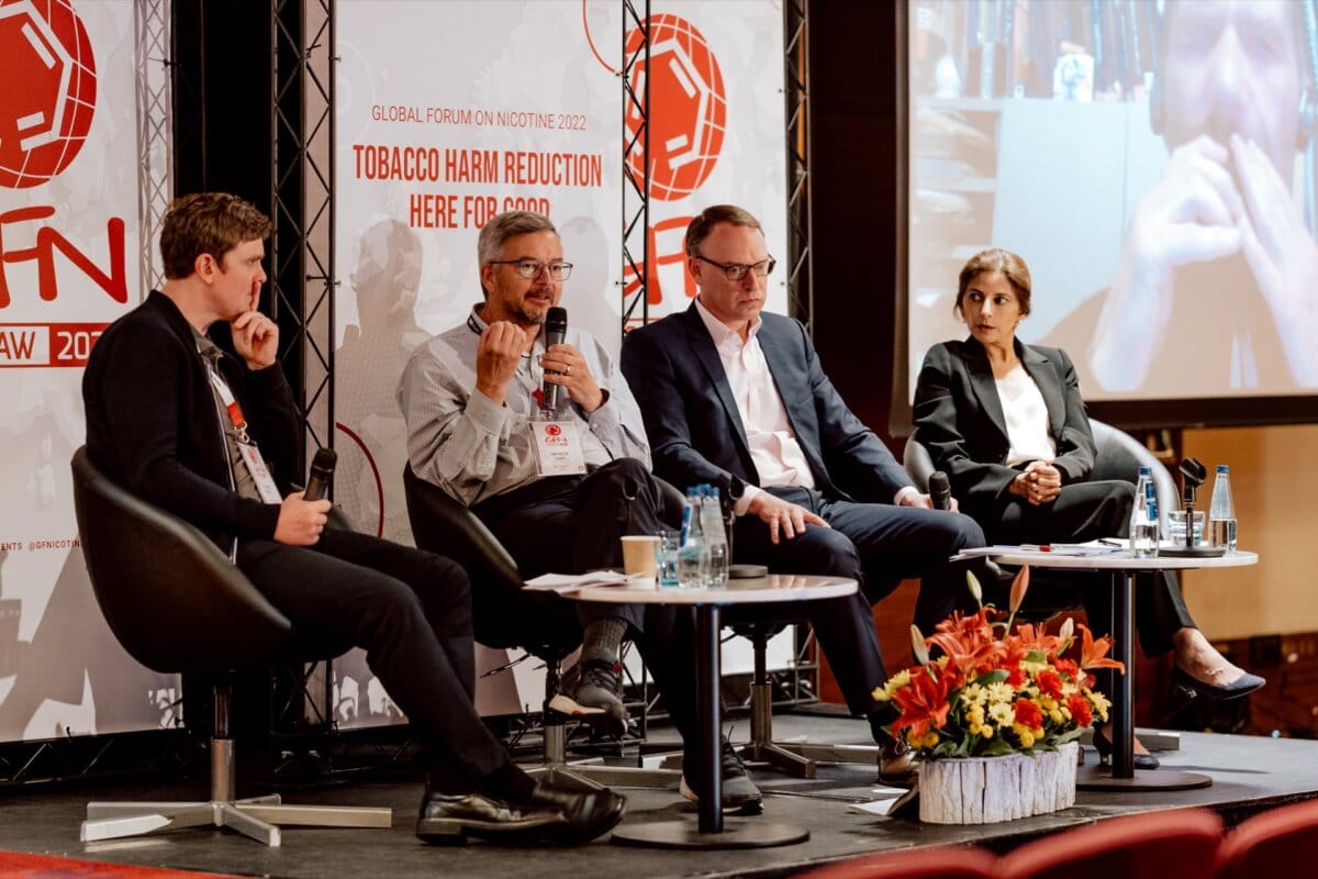 Four people involved in the World Forum on Nicotine 2022 discussion are seated on a stage panel. Two men and a woman are seated on the left in casual business attire, while another man and a woman on the right are in formal attire. Behind them, a video screen displays a fifth participant, perfectly capturing a photo recap of the event.  