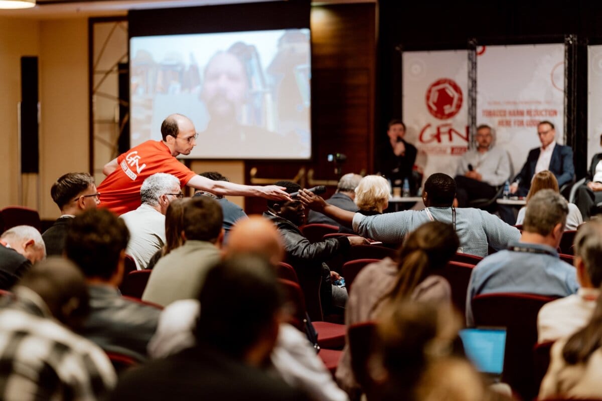 A man in a red shirt stands in the audience and extends his hand to hand a microphone to a participant. The room captured by the event photographer is filled with people sitting in rows facing a panel of speakers and a large screen displaying the person's image. This photo essay of the conference highlights the engaging atmosphere.  