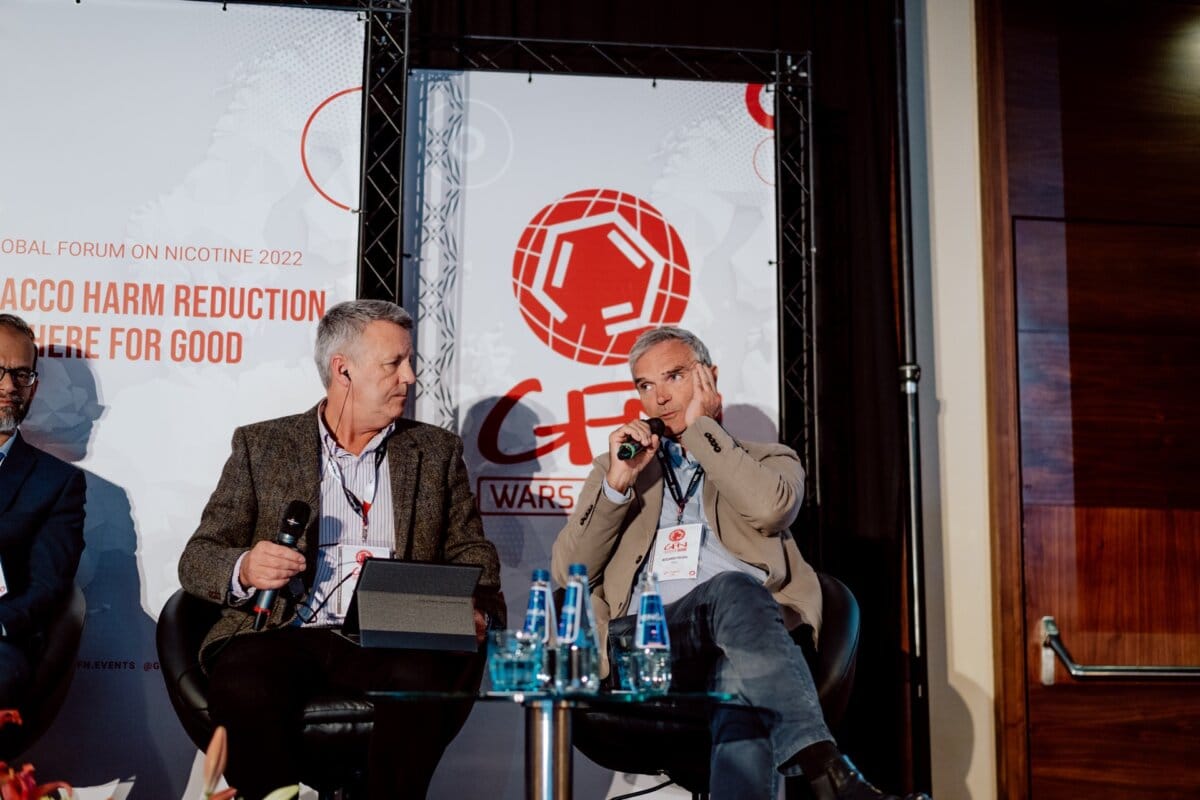 Two men sit on stage during a panel discussion of the World Nicotine Forum 2022 in Warsaw, Poland. They hold microphones, engaging in conversation. In the background are the event's branding and text promoting tobacco harm reduction, creating an eye-catching photo essay of the conference.  