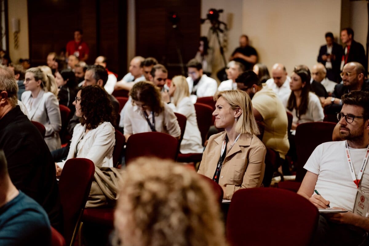 A diverse group of people attentively sitting in rows, some with notepads and lanyards, probably at a conference or seminar. There are red chairs in the room, and several people in the background standing or operating cameras. This is an extensive photo essay of the conference, capturing the essence of the event.  