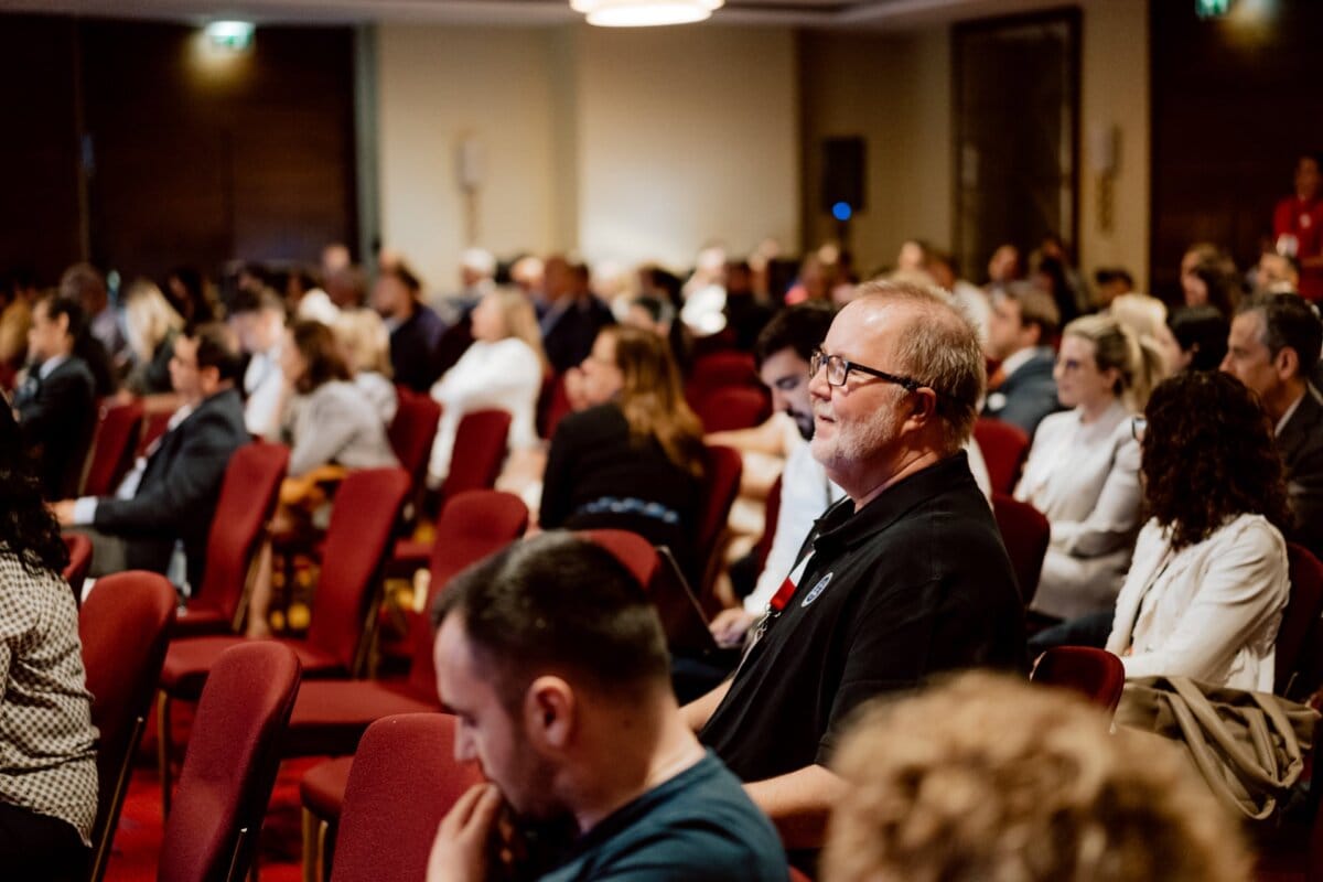 A large group of people sit in rows of red chairs and carefully watch a presentation or speaker in the conference room. The room is dimly lit and most of the attendees are clustered at the front, as captured in a photo report of the conference. The audience seems to be engaged and listening intently.  