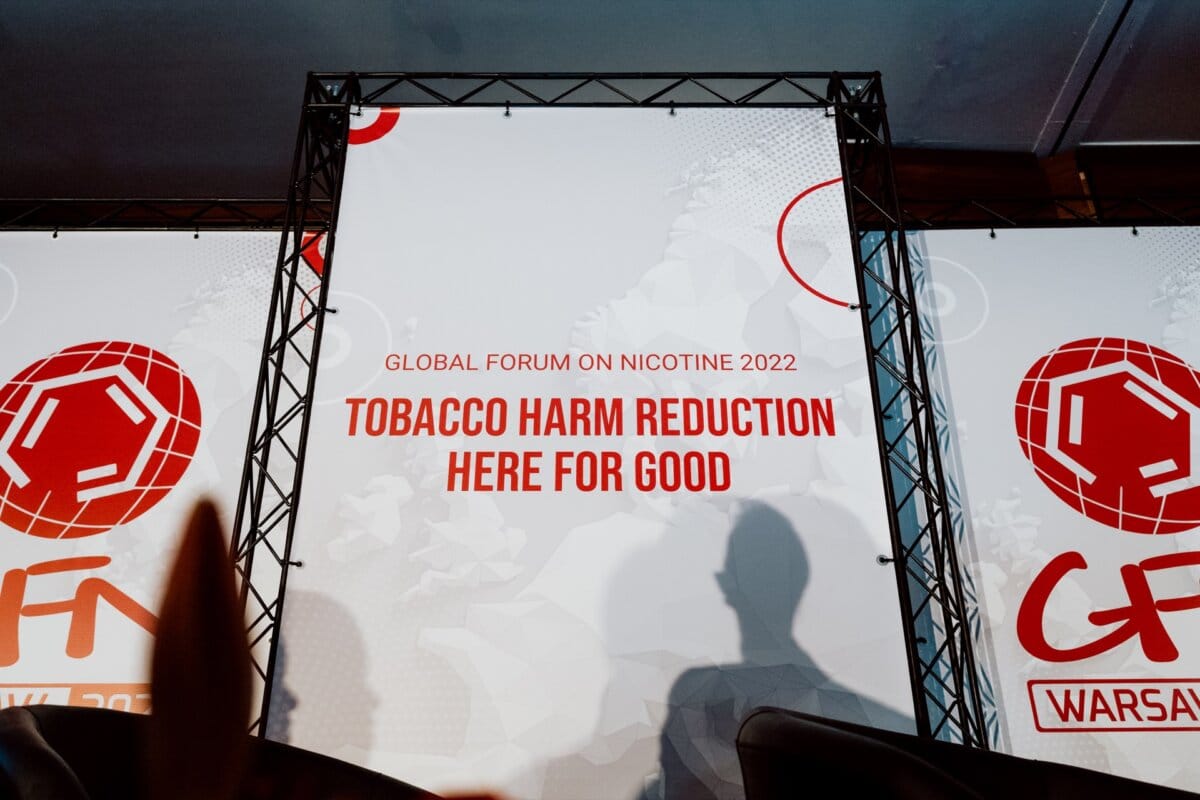 Background image of a scene from the World Forum on Nicotine 2022 with the centrally placed text "Reducing the harm of smoking for good". The design features abstract geometric patterns in red and white, with a visible shadow of a person and additional branding of the conference, perfect for photo coverage of the event. 