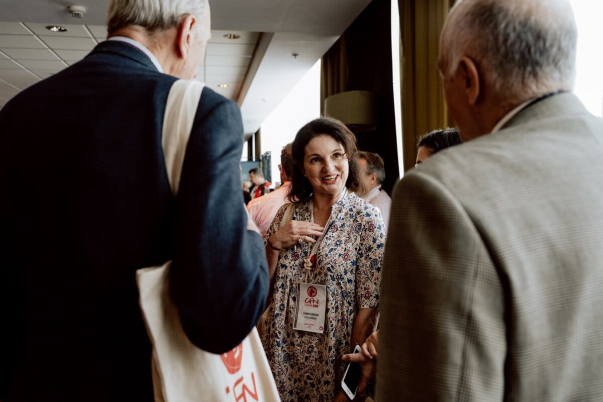 A group of people engaged in conversation at a conference. A woman in a flowery dress with a name badge is talking to two men in suits, one of whom is carrying a bag. The other participants are visible in the background of the brightly lit room, which perfectly captures the essence of the conference photo essay.  