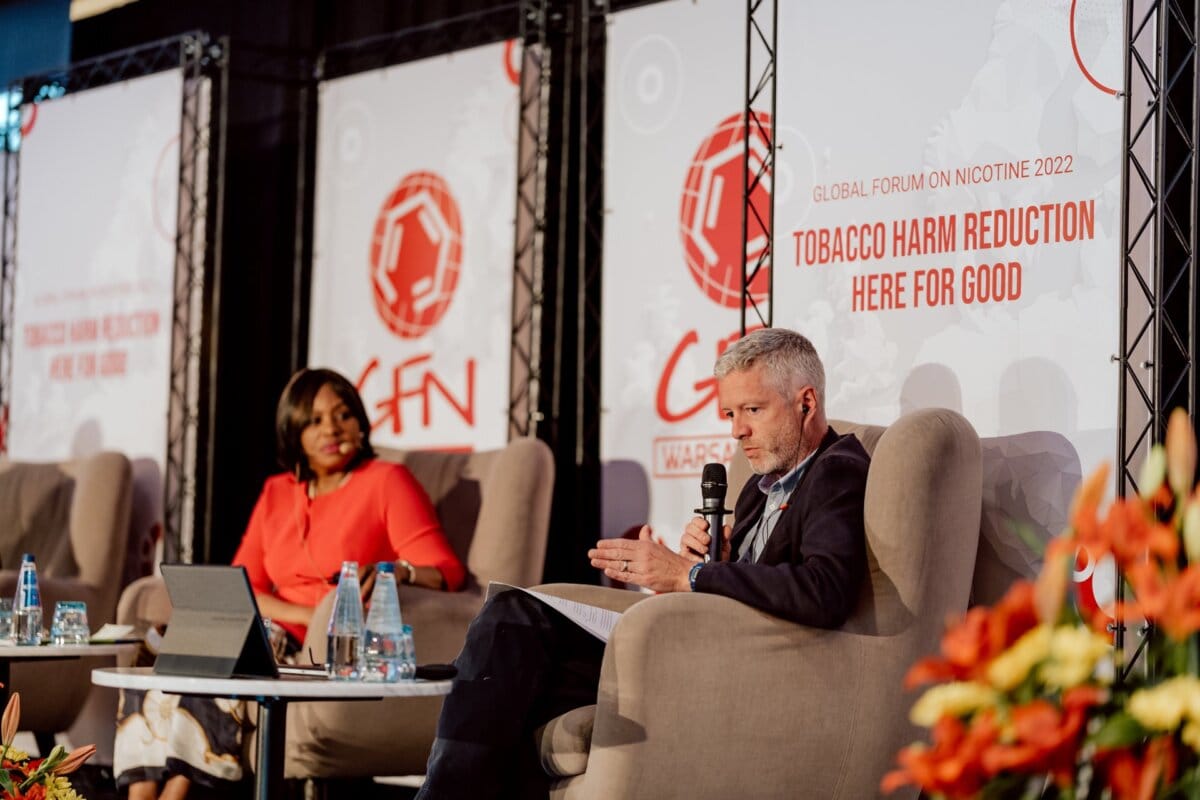 Two people sit on the stage panel of the World Forum on Nicotine 2022. In the background are banners with the slogan of the event: "Tobacco harm reduction: here for good." The two are engaged in a discussion; the man is speaking into a microphone. This event photo shows an engaging snapshot of the conference.   