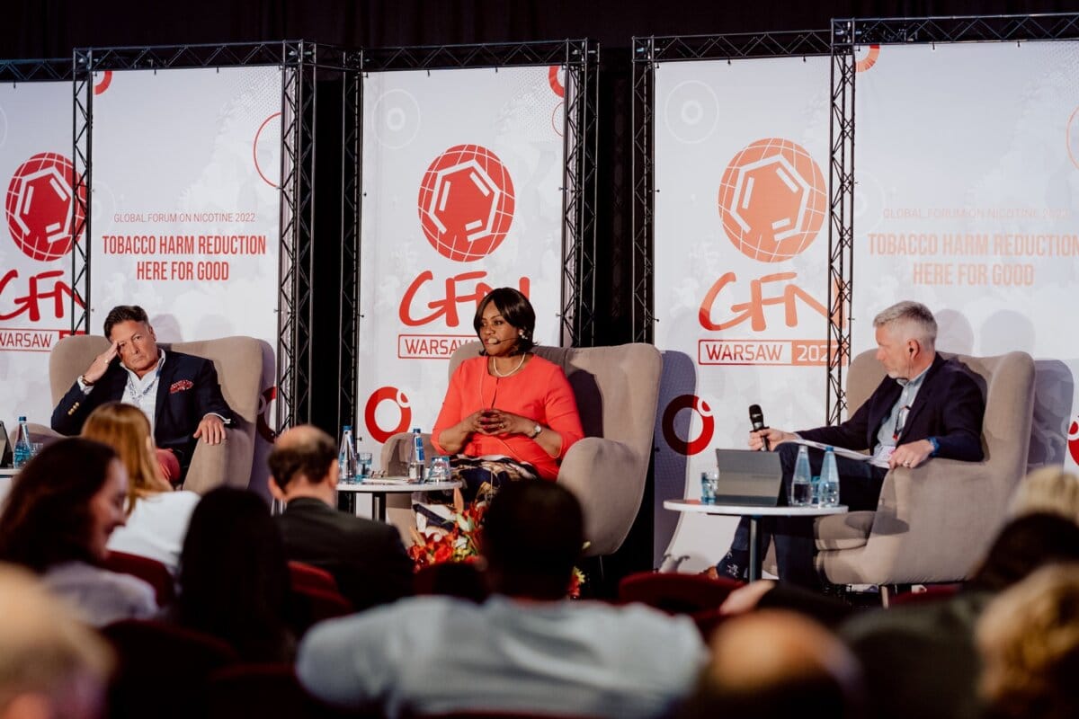 Three people sit on stage during a panel discussion at the World Nicotine Forum 2023 in Warsaw, Poland. Behind them are event banners and a backdrop with the forum logo and the slogan "Reducing tobacco-related harm: here for good." This photo essay from the conference shows the audience visible in the foreground.  