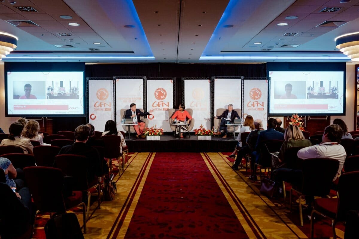 A panel of three sits on a stage facing the audience in the conference room, with the "GFN" logo in the background. Two large screens on either side of the stage show the same image. The audience sits in rows and listens intently as an event photographer captures a detailed photo report of the conference.  