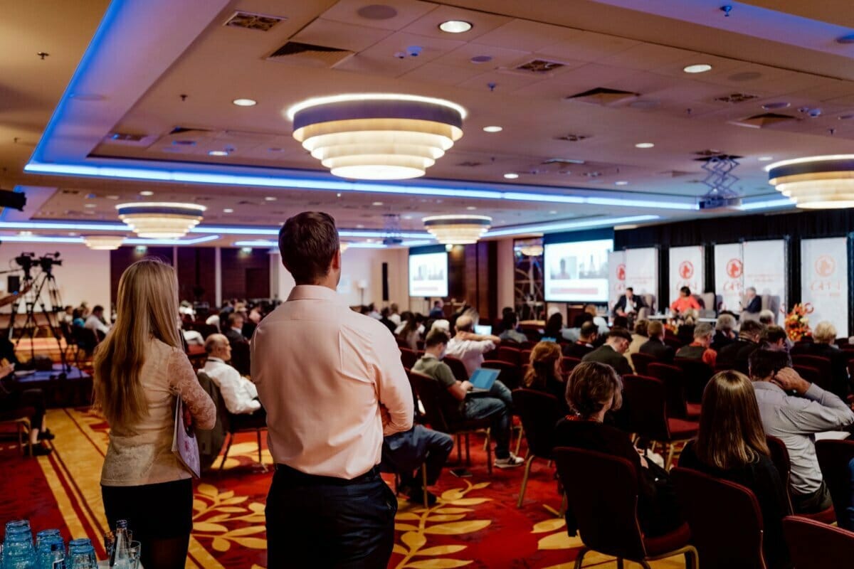 The conference room is filled with attendees sitting in chairs, facing the stage with speakers and presentation screens. The room is warmly lit with modern chandeliers, and two people stand at the back to observe the event as part of a photo tour of the conference. 